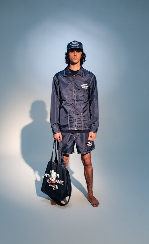 Model standing over seamless background in dark blue with spotlight wearing WHR Hat, Jacket, Shorts, while holding bag.