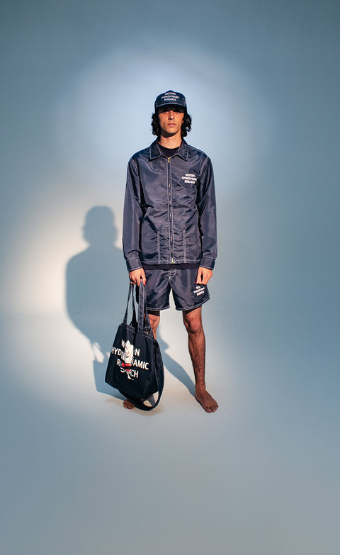 Man wearing WHR x Birdwell collection consisting of a WHR Hat, Competition Jacket, WHR Short and Tote Bag standing over seamless background with spotlight.