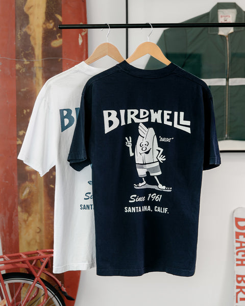 61 Tee in Navy and White on hangers in front of old surfboard and vintage Birdwell jacket in a frame.