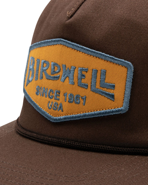 The Service Snapback in Brown, with embroidered gold and blue Birdwell patch and visor rope detail. Close up image.