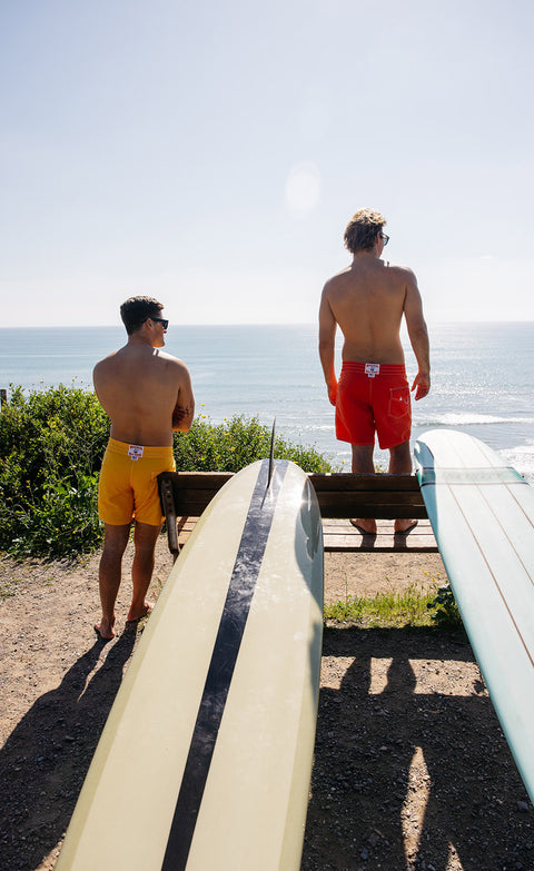 Two guys standing on a cliff on a sunny day wearing the 300 boardshort from Birdwell in yellow and red with longboards leanging on the bench.