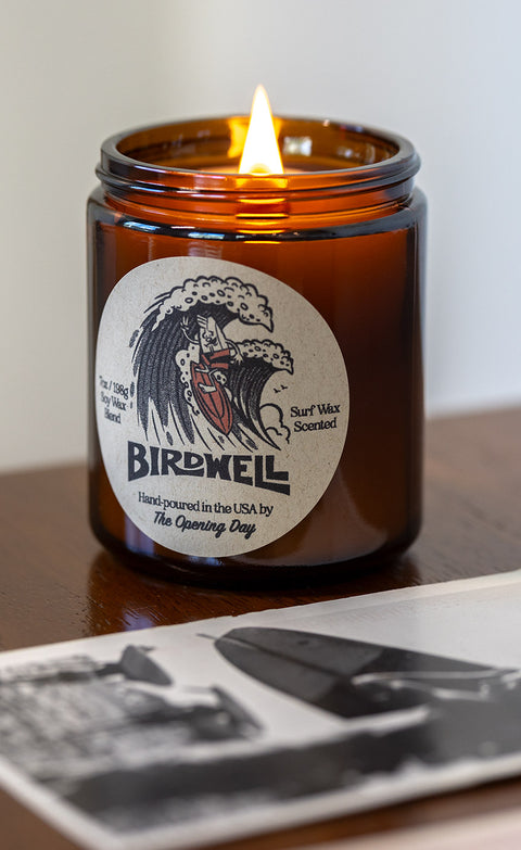 Lit Birdwell Surf Wax Candle on dark wooden desk with black and white photo of surfboard.