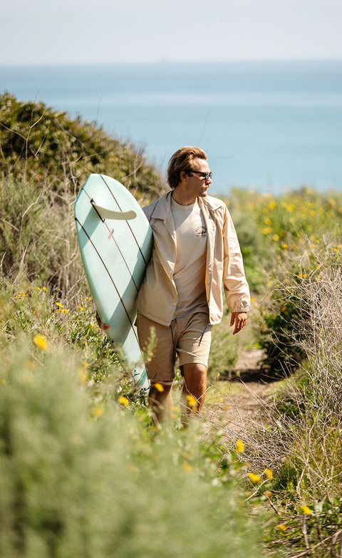 Hillside ocean scenery with man wearing the Tan Coach's Jacket and Tan Drifter Shorts holding a surfboard.