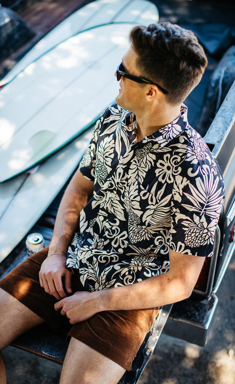 Model wearing the Lanai Shirt in black with brown Drifter Shorts while sitting on truck bed with two surfboards in the background. 