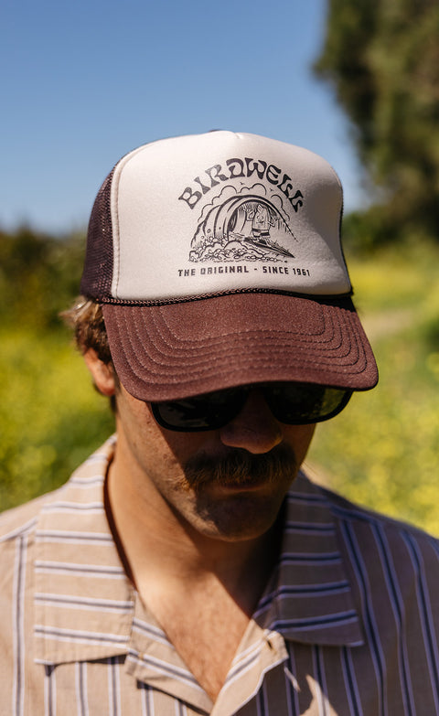 Close up of man wearing the Surfin' Birdie Trucker Hat in Brown wearing a brown striped shirt and sunglasses. Blurred hill of greenery in the background.