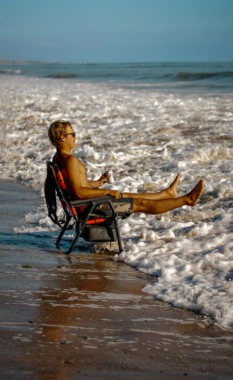 Kevin sitting on Limited-Edition Voyager Chair in black and red with feet up in the air while waves are rolling in on the beach.