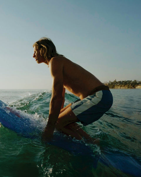Joel Tudor crouches low on his blue longboard while riding a wave, focused ahead, wearing Birdwell 300 Legend boardshorts.