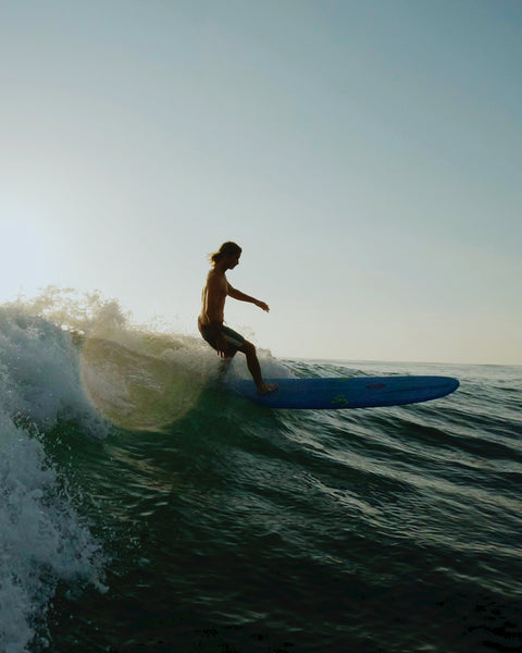 Joel Tudor rides a blue longboard on a wave at sunset, silhouetted against the sky, wearing Birdwell 300 Legend boardshorts.