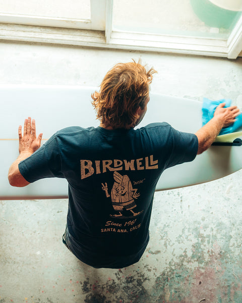 Kevin Skvarna is leaning over a surfboard inside a shaping room, wiping down the surfboard while wearing the 61 t-shirt in navy. The shirt is a Navy T-Shirt over White Background. Cream colored screenprint shows Birdwell logo with birdie holding up a peace sign with 