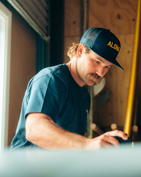 Kevin Skvarna is indoors in front of surfboards and leaning over a surfboard measuring the center. He is wearing the Aloha Hat in Navy which is a trucker hat made out of navy fabric with a black based patch on the front and in gold writing says aloha. 