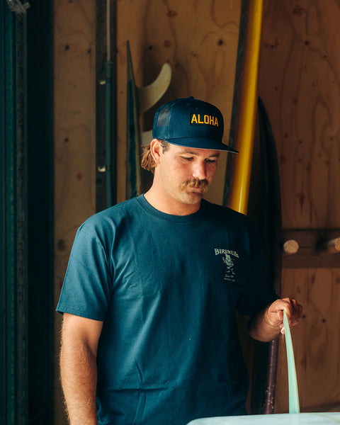 Kevin Skvarna is indoors in front of surfboards and checkin out a fin in a surfboard on a table. He is wearing the Aloha Hat in Navy which is a trucker hat made out of navy fabric with a black based patch on the front and in gold writing says aloha. 