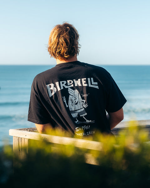 Kevin Skvarna is at a lookout that overlooks the ocean waves and people surfing. In the foreground is green foliage. He is wearing the Birdwell 61 t-shirt in white/black that shows Cream colored screenprint shows Birdwell logo with birdie holding up a peace sign with 