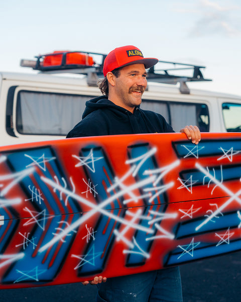 Kevin Skvarna is outdoors in front of a van holding a red, white and blue surfboard. He is wearing the red Aloha trucker hat that has mesh on the back and a black patch with gold lettering that spells ALOHA. 