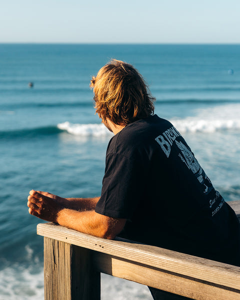 Kevin Skvarna is at a lookout that overlooks the ocean waves and people surfing. He is wearing the Birdwell 61 t-shirt in white/black that shows Cream colored screenprint shows Birdwell logo with birdie holding up a peace sign with 