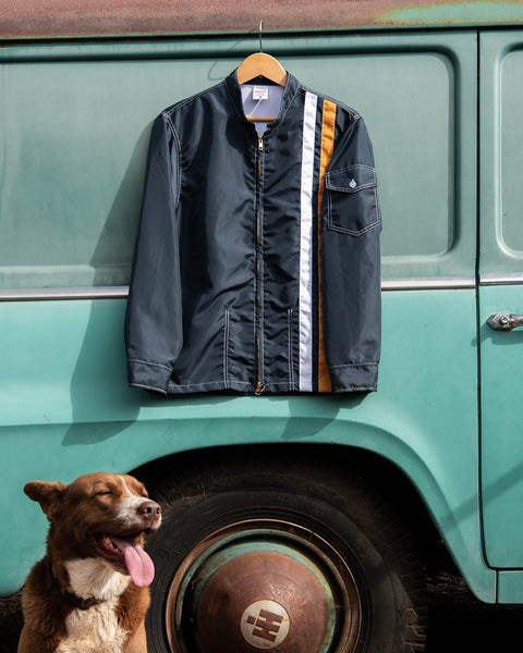 The Birdwell Racing Jacket 25 in Navy with two vertical stripes one white and one yellow hangs on the side of a tourquiose van with a shepherd dog in the foreground. 