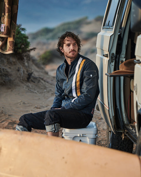 The model is wearing a Navy Racing Jacket with two vertical stripes, one white and one yellow. He is sitting on a cooler outdoors, in front of what looks to be a VW Westfalia van with the door open and a surfboard in the foreground.