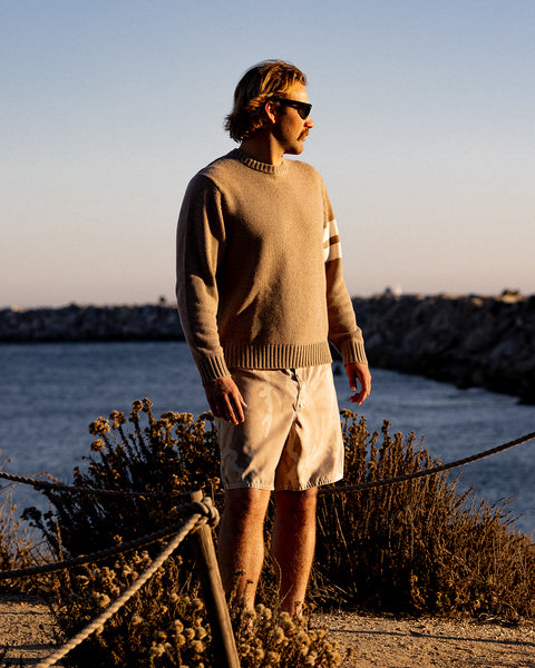Kevin standing by a body of water wearing a bone comp stripe sweater and sunglasses.