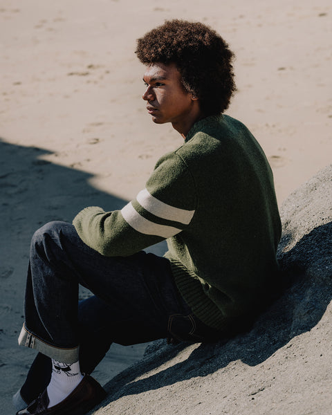 Obi sitting on a sandy surface wearing a dark olive comp stripe sweater with white stripes.