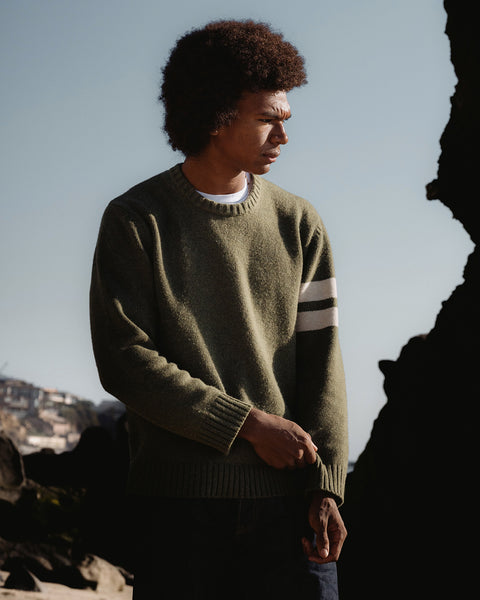 Obi wearing an olive green sweater with a white stripe, standing outdoors against a rocky background.