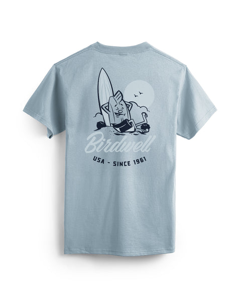 Back View of Arctic Blue T-shirt with a graphic design on the back featuring a Birdie leaning against a surfboard with his foot up on a coconut and text below of Birdwell USA Since 1961. Product on a white background.
