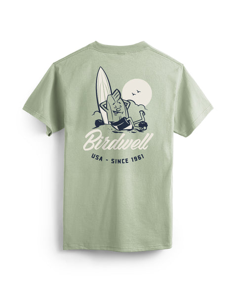 Desert Green T-shirt with a graphic design on the back featuring a Birdie leaning against a surfboard with his foot up on a coconut and text below of Birdwell USA Since 1961. Product on a white background.
