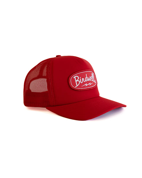 Red trucker hat with a BIRDWELL logo patch and lightning bolt underneath the text on the front of the hat on a white background