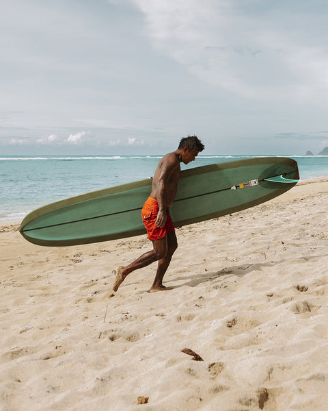 Toots wearing the 300 Boardshorts in Medium Orange, holding a green surfboard under left arm while walking on the sand. Blue ocean shore in background.