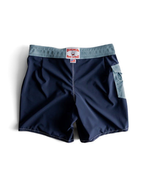 Back view of the 808 boardshorts, navy body with federal blue waistband and pocket. White License plate label with Made in USA Label beneath. Product on white background.