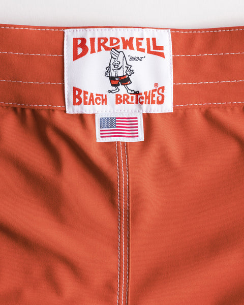 Close-up of the 808 SurfStretch Side Pocket Boardshorts in Paprika, featuring vibrant orange SurfStretch® fabric with white stitching, a Birdwell Beach Britches label with their signature cartoon character, and a small American flag tag—celebrating the classic boardshort style made in the USA.