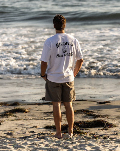 Rear view of a person standing barefoot on sandy beach, hands in pockets, wearing the Birdwell License Plate T-Shirt in white with black print, facing the shoreline as small waves roll in.