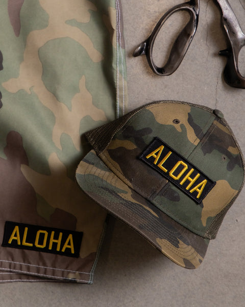 Camouflage boardshorts and hat with aloha patches on a cement background. Scissors in the upper right corner.
