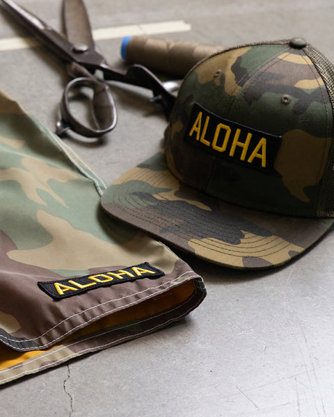 Camouflage hat and boardshorts with aloha patches on a cement surface with scissors.