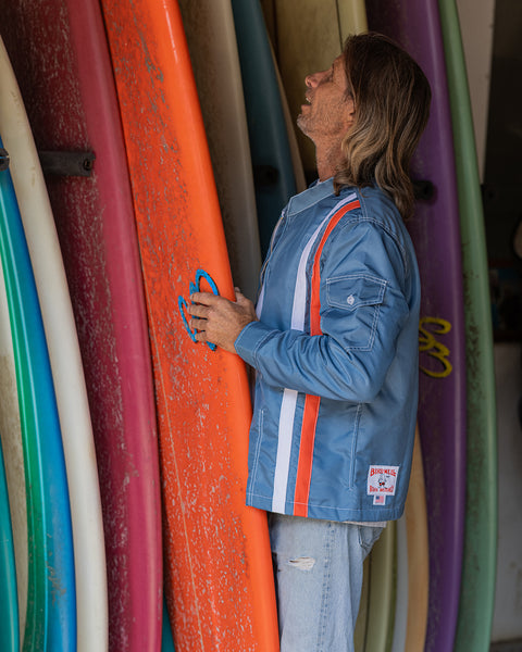 Side view of Joel Tudor inspecting a vibrant orange surfboard while standing in front of a rack of surfboards. He’s wearing the Birdwell Racing Jacket 25 – Le Mans, with its distinctive vertical orange and white stripes running down the sleeve and side panel.