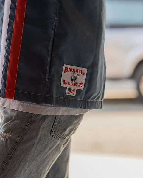 Close-up of the lower side of the Birdwell Racing Jacket 25 – Le Mans, highlighting the orange and white racing stripes, the Birdwell Beach Britches logo, and the American flag patch that reads “Made in U.S.A.” Joel’s distressed light blue jeans and layered white t-shirt are also visible.