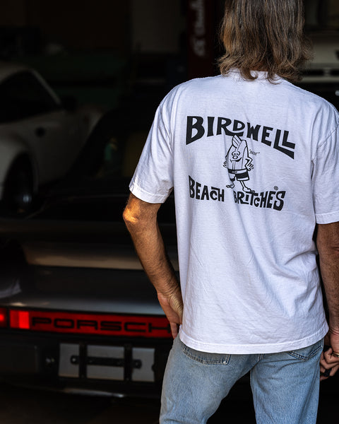 Rear view of a person wearing the Birdwell License Plate T-Shirt in white with black print and light blue jeans, standing in front of a classic Porsche inside a dimly lit garage.