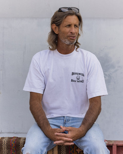 Person seated on a patterned bench cushion wearing the Birdwell License Plate T-Shirt in white with black print and light blue jeans, sunglasses resting on their head, looking off to the side with a relaxed expression.