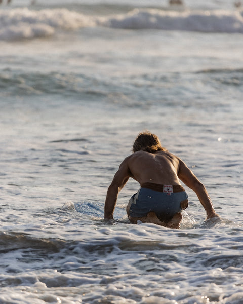 Joel Tudor kneels on his blue longboard in shallow water with waves around him, wearing Birdwell 300 Legend boardshorts.