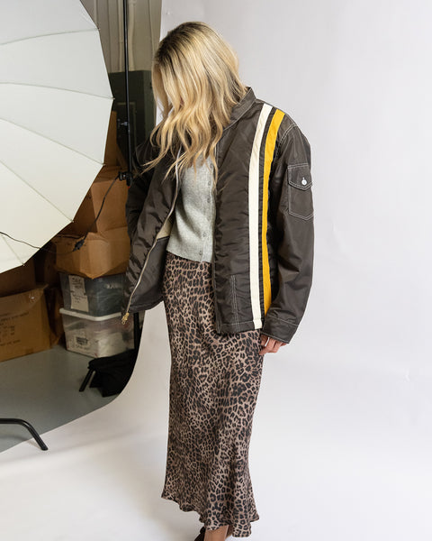 Birdwell Quilted Racing SurfNyl® jacket in brown on a woman model taken from the left side. The jacket has a pocket on the left arm with yellow and white stripe  accents going down the front of the jacket, and wearing a patterned skirt on a white background