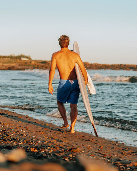 Man walking on a beach with a surfboard, backlit by the sun.