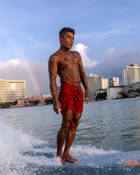 Man in red swim trunks standing in water with a city skyline and rainbow in the background