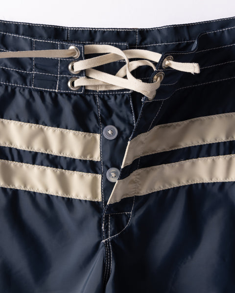 Navy blue swim shorts with beige stripes and white laces on a light gray background
