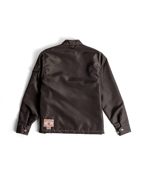 The Racing Jacket 26 - Brown/Bone/Gold is laid flat, back up, on a white background. It features a dark brown color, collared design, visible stitching, buttoned cuffs, and a rectangular Birkenhill patch near the lower left hem.