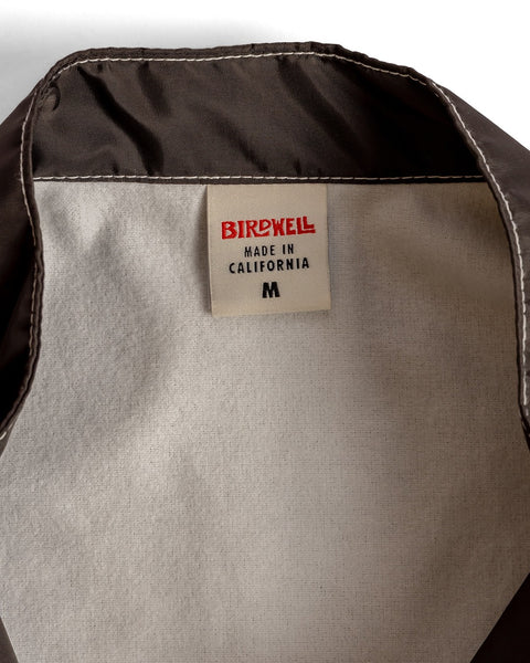 Close-up of a clothing tag inside a brown Racing Jacket 26 - Brown/Bone/Gold. The tag reads “BIRDWELL MADE IN CALIFORNIA” with a bold red logo, plus a black “M” indicating the medium size of this American-made jacket.