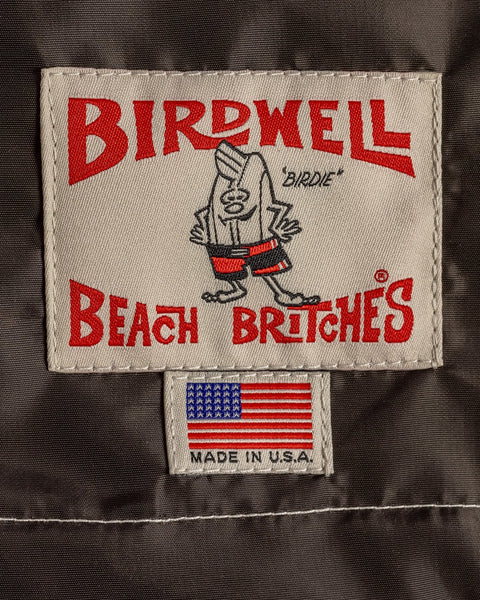 A fabric label on the Racing Jacket 26 - Brown/Bone/Gold shows a cartoon bird in shorts, Birdwell Beach Britches text, plus a U.S. flag tag with Made in U.S.A.—highlighting its classic SurfNyl and American-made quality.
