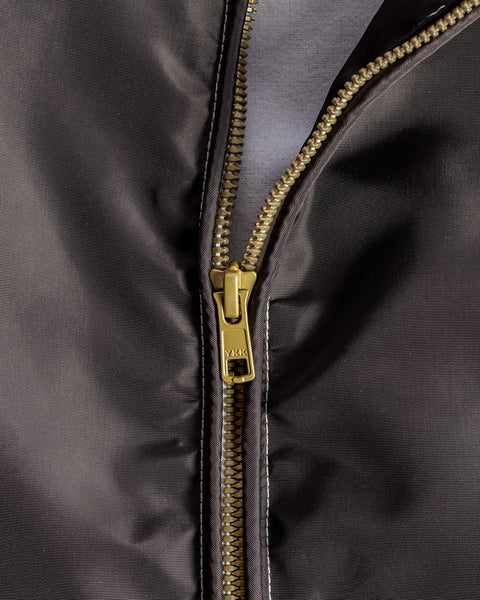 A close-up of a partially unzipped brass YKK zipper on dark, shiny SurfNyl fabric from the Racing Jacket 26 - Brown/Bone/Gold.