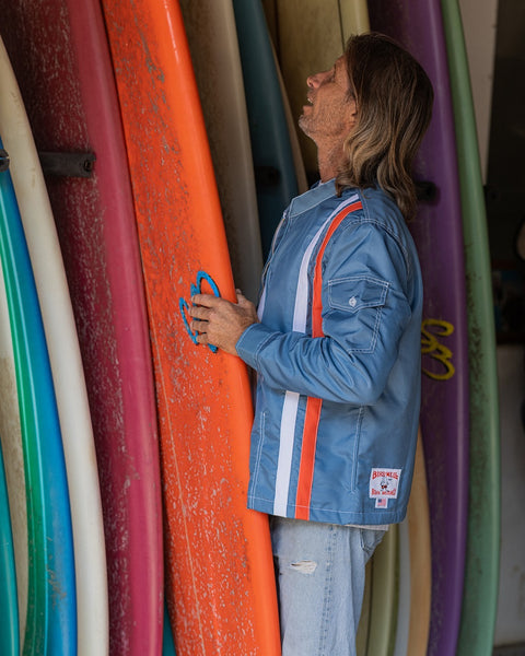 A man wearing a Racing Jacket 26 in Federal Blue/White/Orange stands by colorful surfboards, looking up with his hand resting on an orange SurfNyl surfboard.