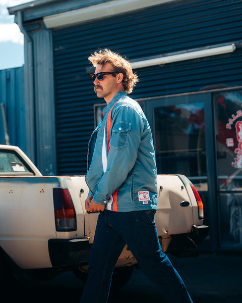 A man with blond hair and a mustache, wearing sunglasses, jeans, and a Racing Jacket 26 - Federal Blue/White/Orange, walks past a white pickup truck in front of a blue industrial building.