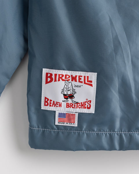 A close-up of the Racing Jacket 26 in Federal Blue/White/Orange reveals a white patch with red Birdwell Beach Britches text and a cartoon bird, plus a tag showing the American flag and “Made in U.S.A.,” emphasizing its American-made quality.