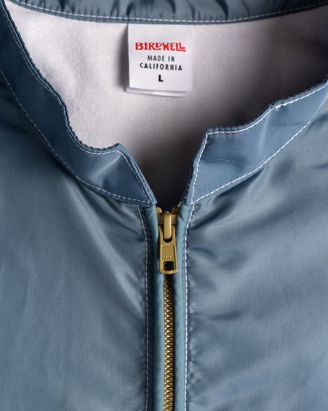 Close-up of the Racing Jacket 26 in Federal Blue/White/Orange, made in California. A gold zipper is partially unzipped, revealing a BIRDWELL MADE IN CALIFORNIA L tag. White stitching outlines the collar of this vintage-inspired jacket.