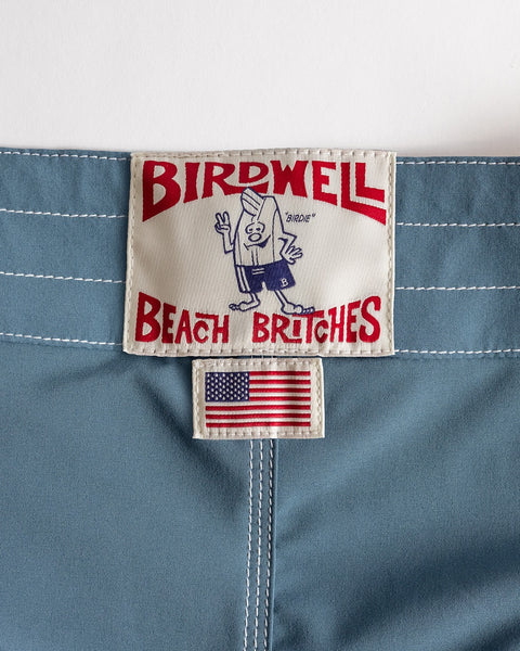 A close-up of the 808 Racing Stripes boardshort in Federal Blue/Bone, showing two sewn labels: one with BIRDWELL BEACH BRITCHES and a cartoon, another below with the American flag—proudly made in USA.